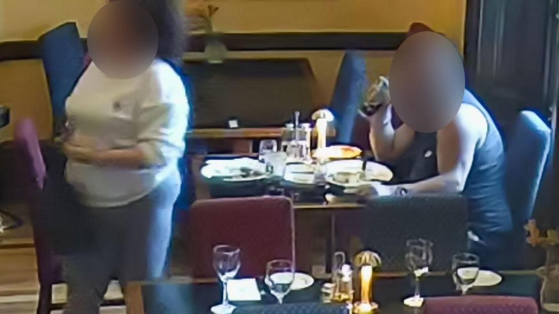 A man in a vest sat at a table with empty plates in front of him. He is drinking from a pint glass of coke. His face is blurred. On the other side of the table, a woman in grey leggings and a white sweatshirt is stood. Her face is also blurred. There are other restaurant tbales set with plates, cutlery and glasses around them.