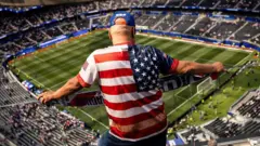 アメリカ国旗のTシャツを着た男性が、サッカースタジアムの上層席からコートを見下ろしている