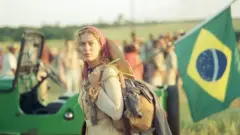 A imagem mostra uma mulher em um cenário rural, com expressão séria e determinada, segurando um facão enquanto caminha à frente de uma bandeira do Brasil. Vestida com roupas simples e um lenço na cabeça, ela tem ao fundo uma paisagem de campos abertos e um veículo carregado, em uma cena que remete à luta pela terra e aos conflitos no campo.