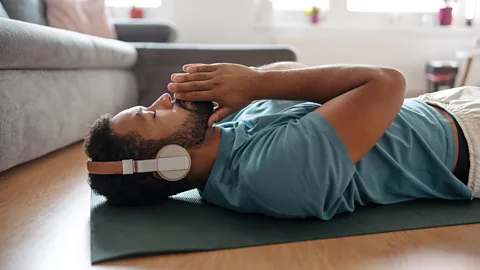 A man lies on a yoga mat on the floor wearing a pair of headphones (Credit: Getty Images)
