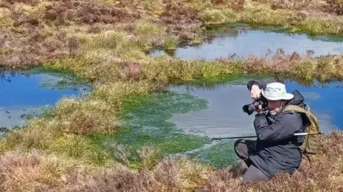 A man kneels on uneven, grassy terrain surrounded by shallow pools of still water. The person is dressed in outdoor clothing, including a hat and backpack, and is holding a camera up to their face, aiming toward the ground or nearby water. The landscape is open and expansive, covered with brown and green vegetation typical of moorland. Small reflective pools of water are scattered across the scene, some edged with bright green algae. In the background, rolling hills stretch across the horizon under a sky filled with soft, layered clouds.